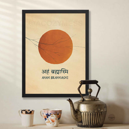 A framed art print featuring a large orange circle on a beige background with the text 'AHAM BRAHMASI' and Devanagari script, hanging on a wall above a table with a metal teapot and two small cups.