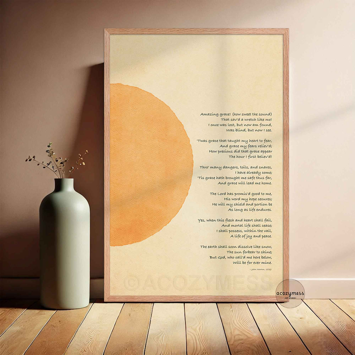 A framed art print featuring a christian hymn with a large abstract orange circle on a cream background, leaning against a wall next to a ceramic vase on a wooden floor.