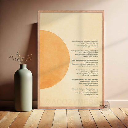 A framed art print featuring a christian hymn with a large abstract orange circle on a cream background, leaning against a wall next to a ceramic vase on a wooden floor.