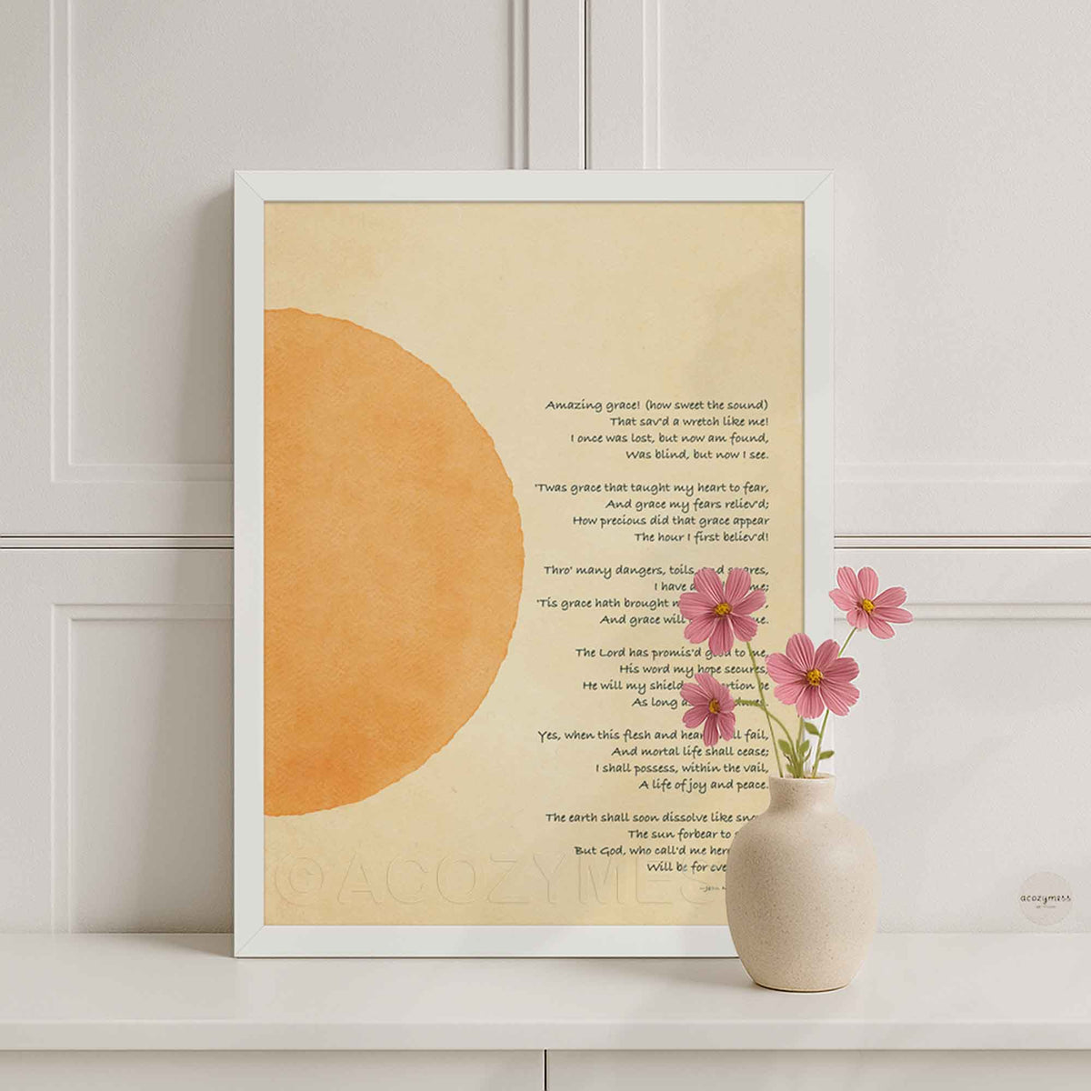 A white framed art print featuring a minimalist design with a large warm orange abstract sun on a cream background and printed christian hymn - amazing grace , displayed leaning against a white paneled wall on a white shelf.