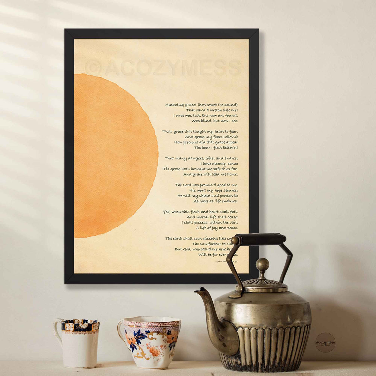 A framed art print featuring the lyrics to 'Amazing Grace' in black text over a beige background with a large orange abstract shape, displayed on a wall above a table.