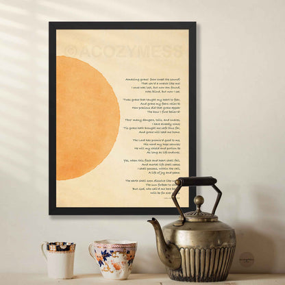 A framed art print featuring the lyrics to 'Amazing Grace' in black text over a beige background with a large orange abstract shape, displayed on a wall above a table.