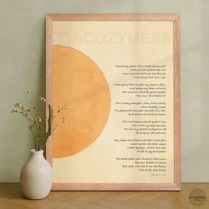 A framed art print featuring a poem titled 'Amazing Grace' with an abstract orange watercolor wash on a cream background, styled in a wooden frame on a wooden surface against a sage green wall.