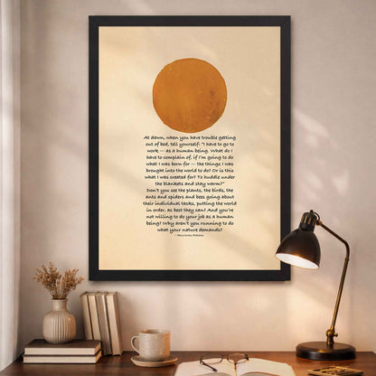 Black Framed artwork with a sun illustration and Marcus Aurelius quote  on a wall above a desk .