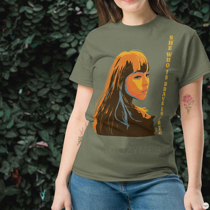 A woman wearing military green t-shirt featuring an illustration of a woman and the quote, She who is brave is free, inspired by Seneca. The design reflects strength and elegance, making it perfect for casual and versatile wear.