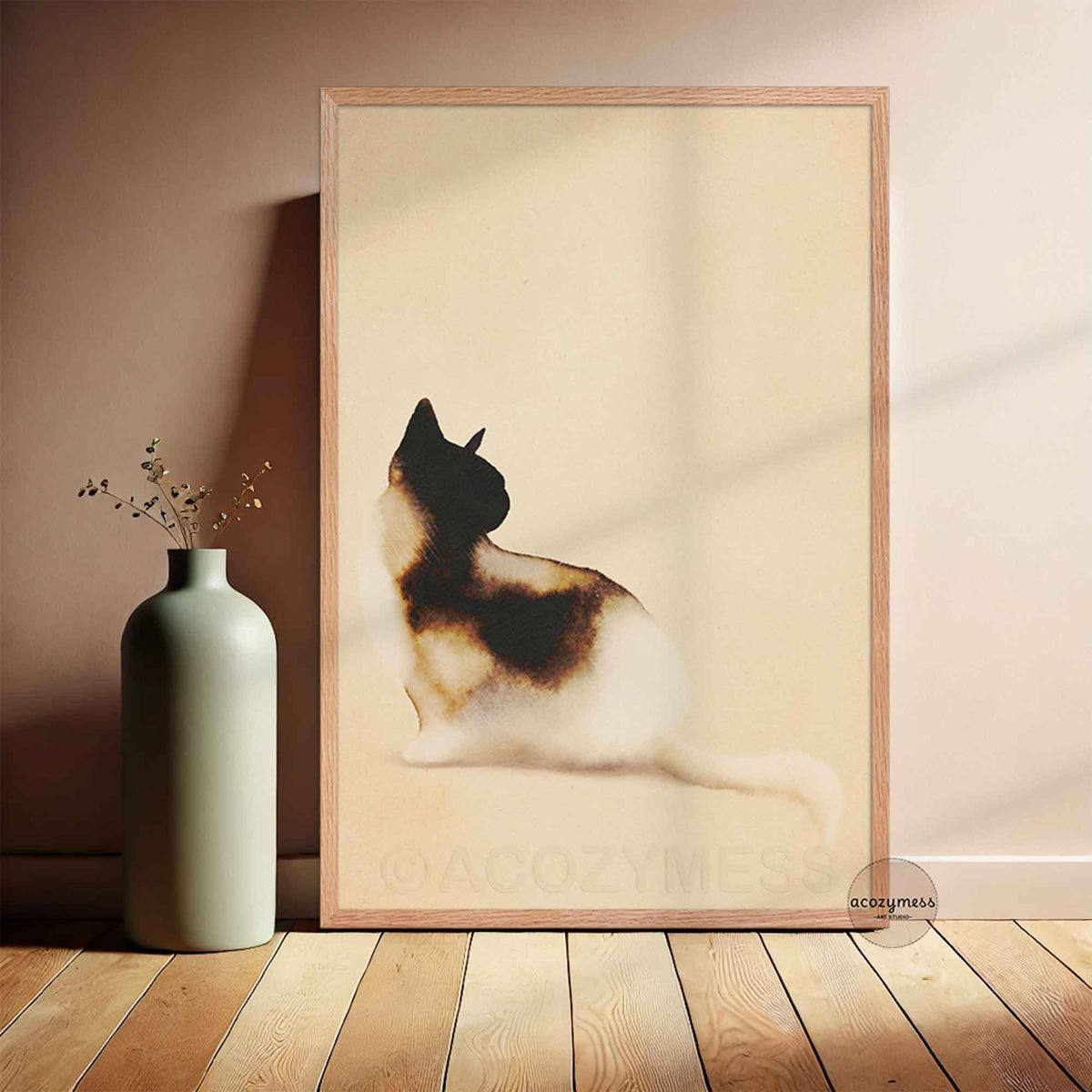 Minimalist watercolor calico cat art poster in a light oak frame leaning against a warm beige wall beside a sage green vase