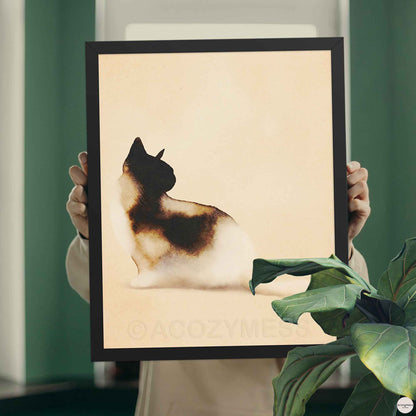 Hands holding a black framed calico cat watercolor print against a green wall with a plant