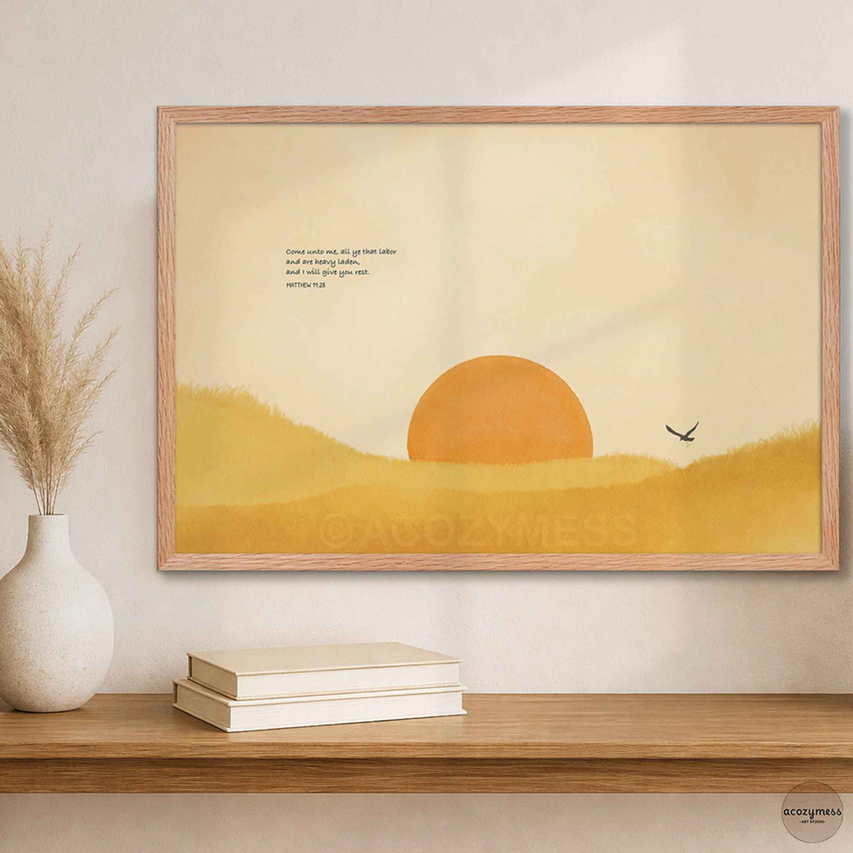 Oak framed Bible verse sun landscape print above a wooden shelf with books and a white vase of dried grass