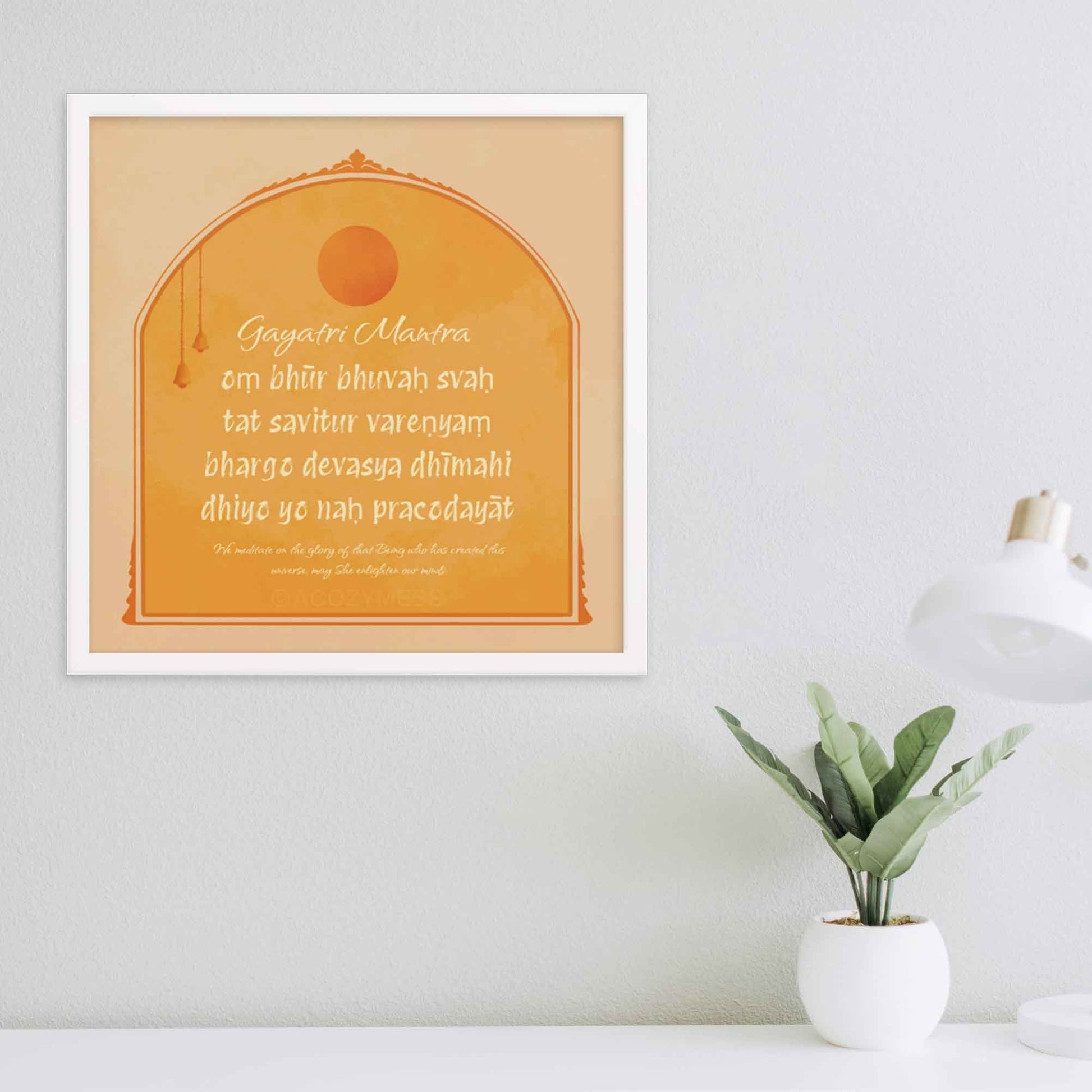 "Gayatri Mantra art print in yellow, beige, and orange colors, designed in square sizes to bring warmth and spiritual energy to any space." in white frame.