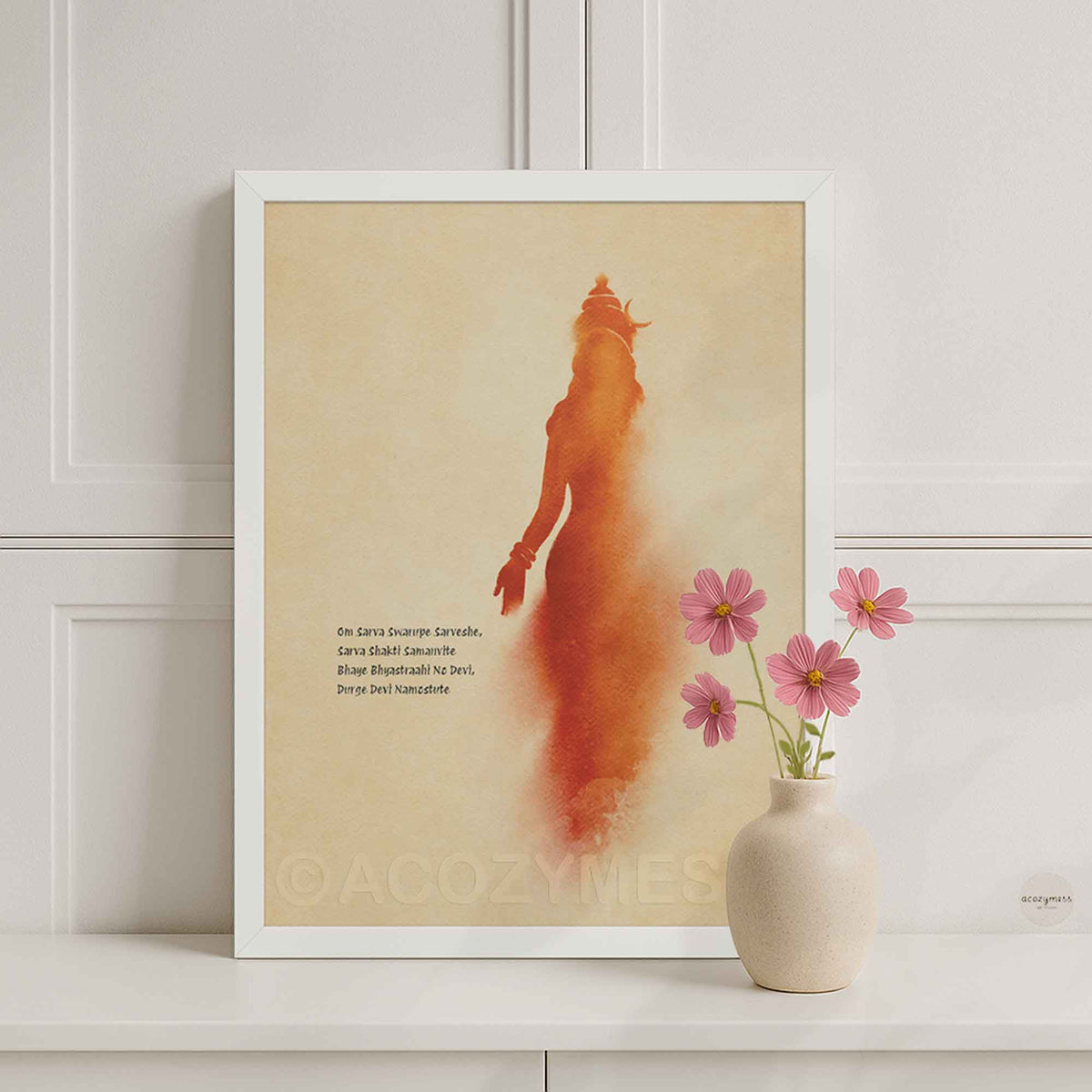 A white framed art print featuring a stylized orange-red silhouette of goddess on a warm beige background, styled on a white shelf next to a small vase with pink flowers against a white paneled wall.