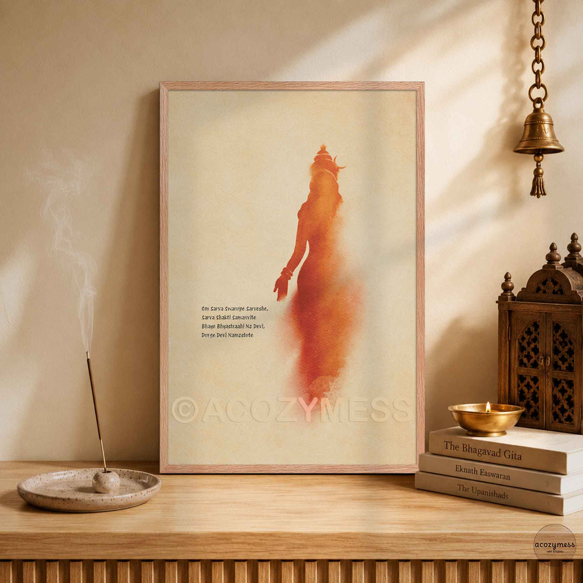 A wood framed art print featuring a stylized orange-red silhouette of goddess durga with mantra on a warm beige background, displayed in a temple.