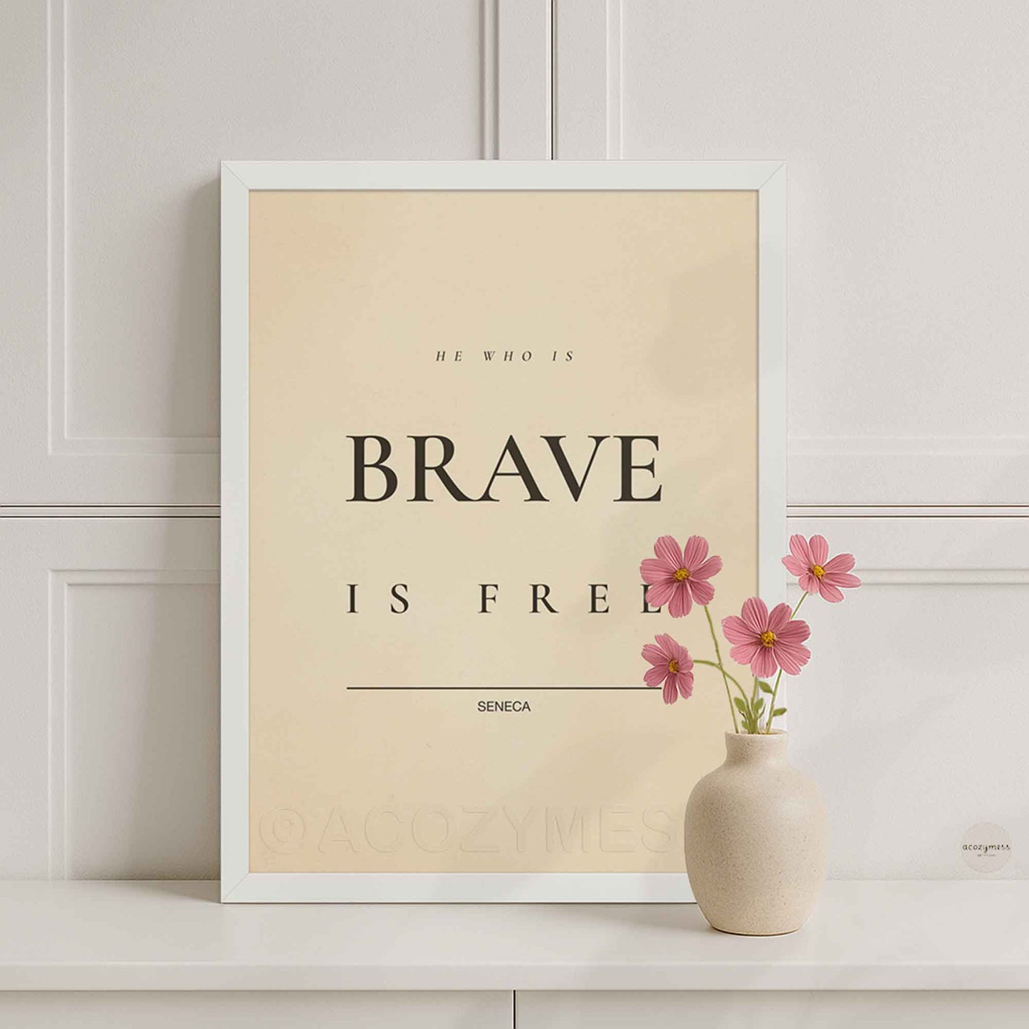 White Framed motivational poster with 'He who is brave is free' quote by Seneca, placed on a shelf with a vase of pink flowers.