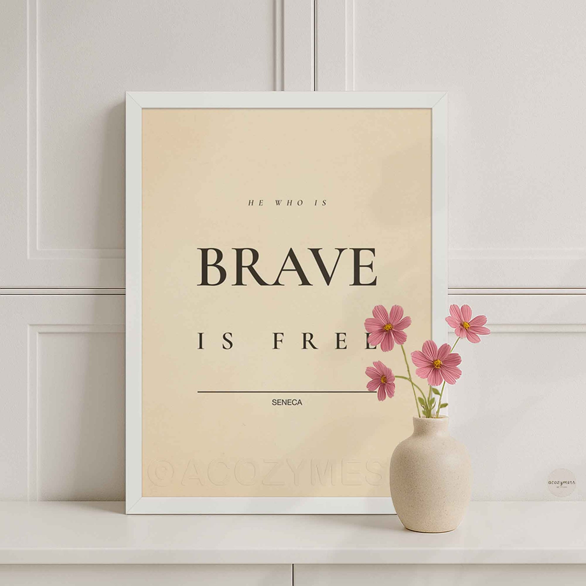 White Framed motivational poster with 'He who is brave is free' quote by Seneca, placed on a shelf with a vase of pink flowers.