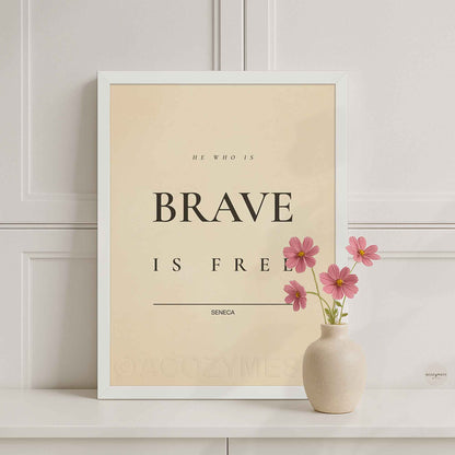 White Framed motivational poster with 'He who is brave is free' quote by Seneca, placed on a shelf with a vase of pink flowers.