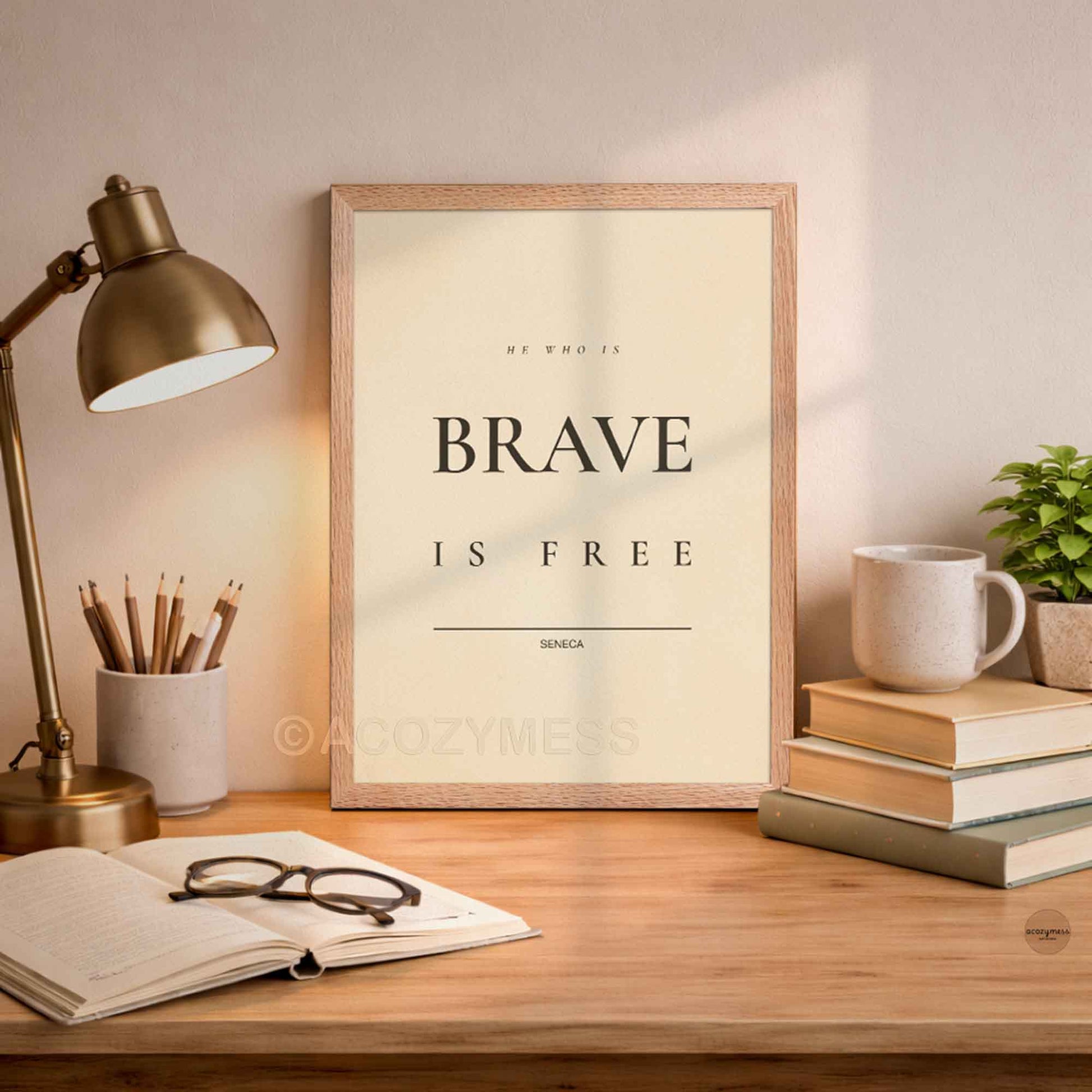 Oakwood Framed print with 'He who is brave is free' quote on a desk with lamp, books, and mug.
