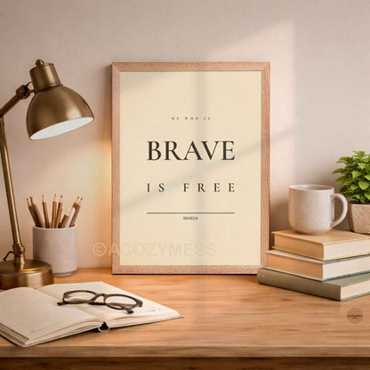 Oakwood Framed print with 'He who is brave is free' quote on a desk with lamp, books, and mug.
