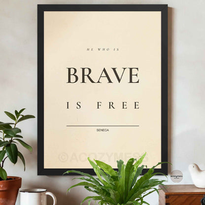 Black Framed poster with 'He who is brave is free' quote by Seneca on a wall with plants and a mug.