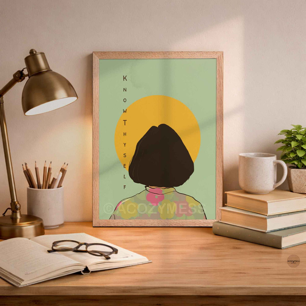 A framed art print featuring a minimalist illustration of a person with a short dark bob haircut seen from behind, standing in front of a yellow sun on a pale green background. The print is displayed on a wooden desk in a styled room setting with a brass desk lamp, an open notebook, glasses, stacked books, and a mug.