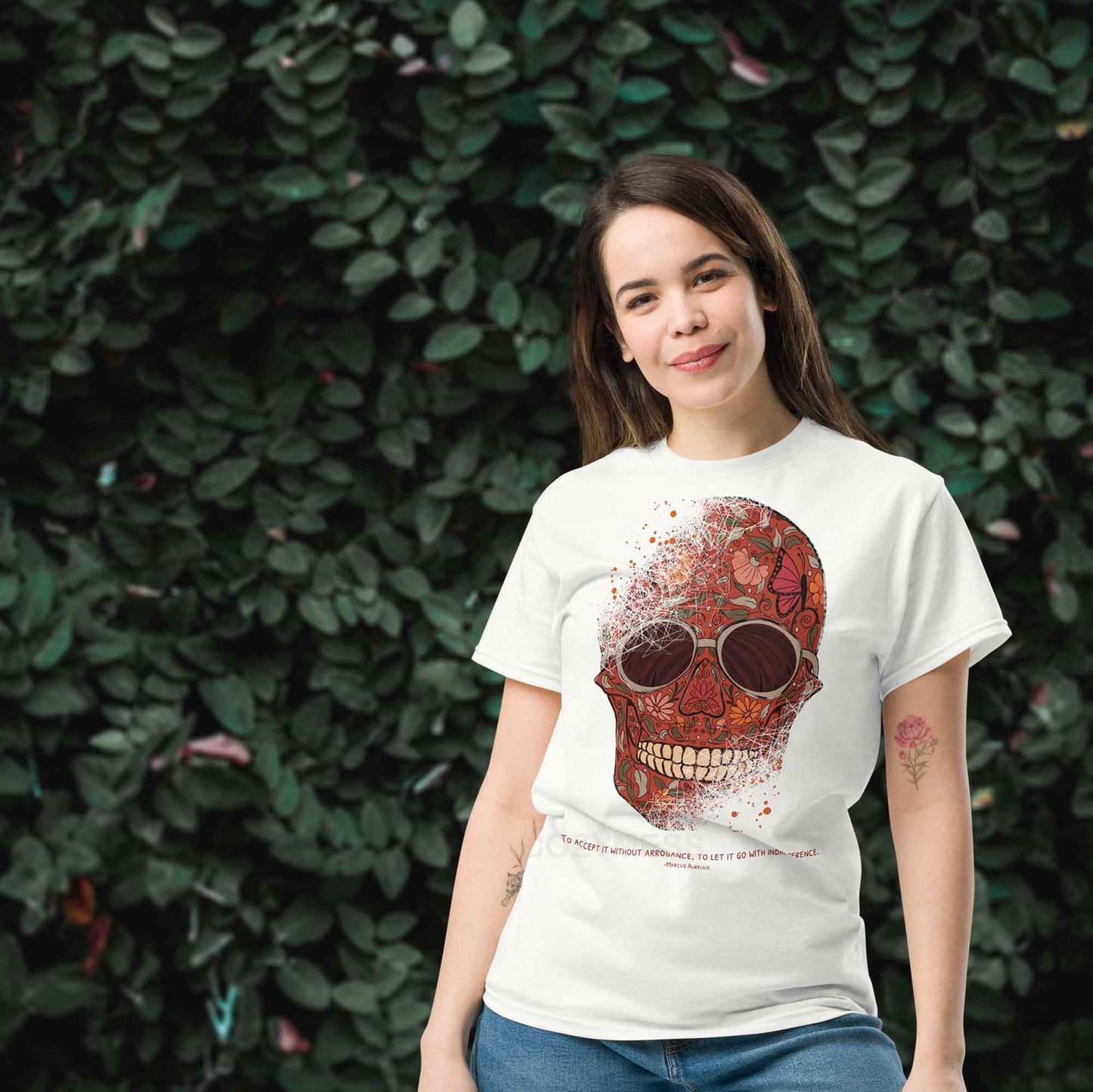 A woman wearing white T-shirt featuring a detailed skull and floral design, paired with a Marcus Aurelius quote: "To accept it without arrogance, to let it go with indifference." The artwork symbolizes memento mori and Stoic philosophy, blending mortality with beauty.