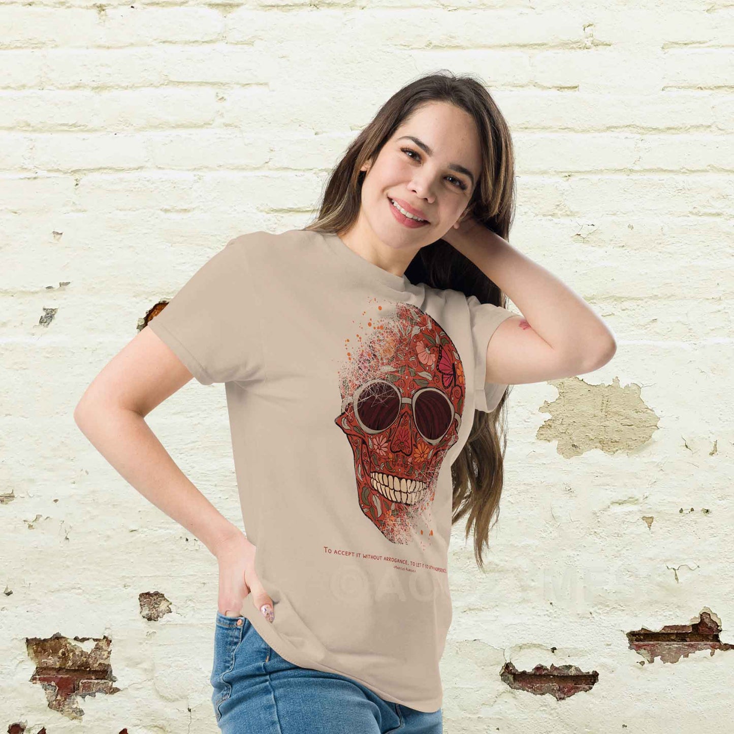 A woman wearing sand color T-shirt featuring a detailed skull and floral design, paired with a Marcus Aurelius quote: "To accept it without arrogance, to let it go with indifference." The artwork symbolizes memento mori and Stoic philosophy, blending mortality with beauty.