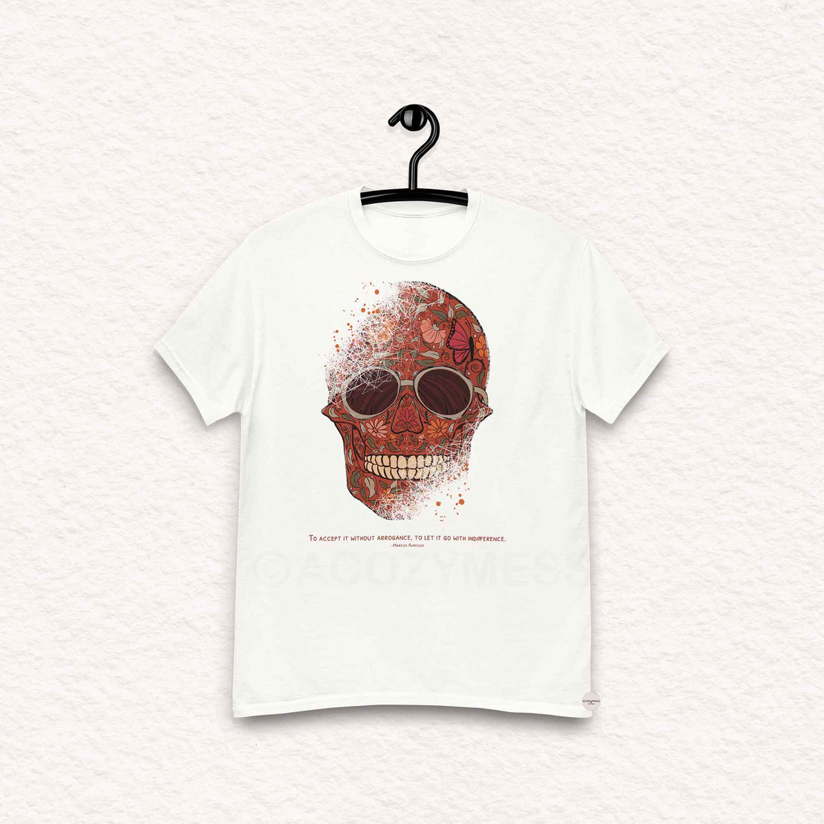 A white  Tshirt featuring a detailed skull and floral design, paired with a Marcus Aurelius quote: "To accept it without arrogance, to let it go with indifference." The artwork symbolizes memento mori and Stoic philosophy, blending mortality with beauty.