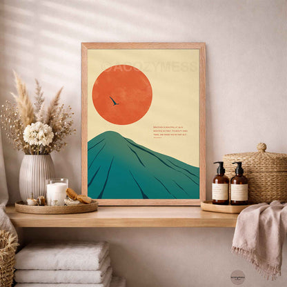 A framed stoic minimalist art print featuring a large orange sun and teal mountain on a cream background, styled on a wooden shelf in a warm, neutral-toned room with dried flowers, candles, and woven decor.