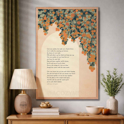 A framed wall art print featuring a poem on marriage with a peach background and a tree made of orange and teal leaves, displayed in a styled room above a wooden console table.