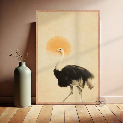 A framed art print featuring a black and white ostrich walking in front of a warm orange sun on a beige background, styled in a minimalist room.