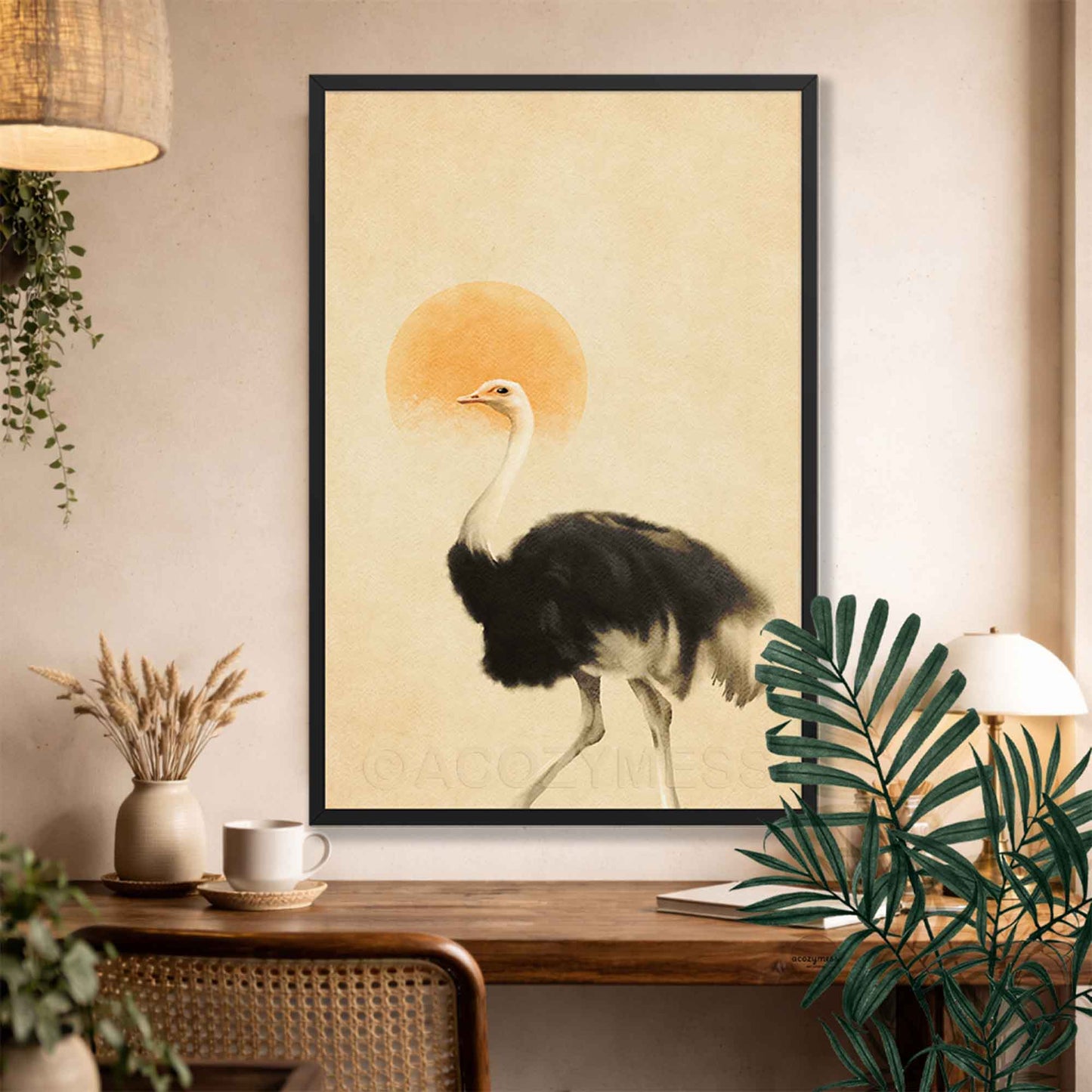 A framed art print featuring a black and white ostrich walking in front of a warm orange sun on a beige background, displayed on a wall above a wooden console table with plants and home decor.