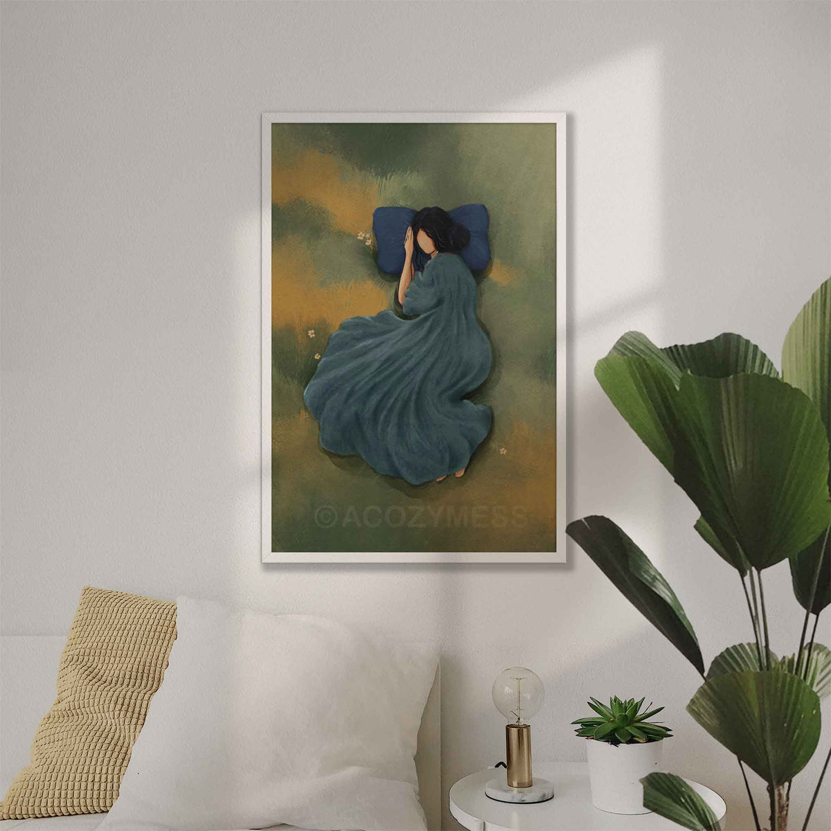 Minimalist white framed art poster of a woman sleeping peacefully in a green grassy meadow with a blue pillow, styled in a bright, calm bedroom wall.