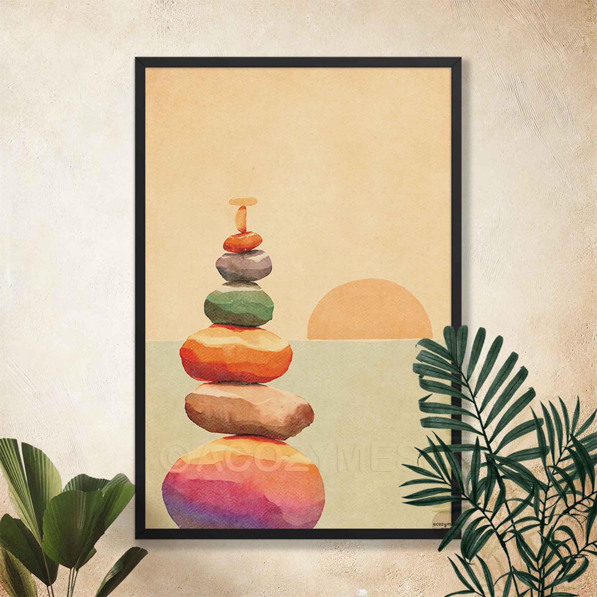 A framed art print featuring a stack of colorful stacked stones and a sun on a textured beige background, styled on a wall with green tropical leaves.