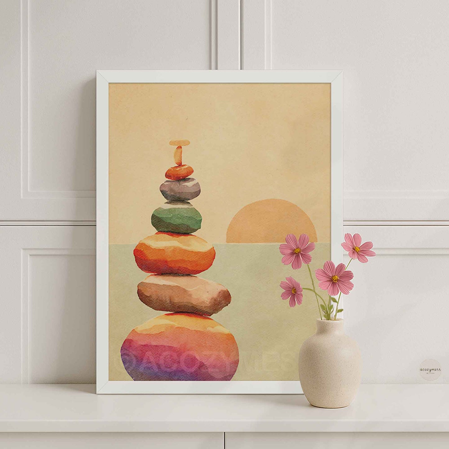A white framed art print featuring a stack of colorful zen stones and a sun on a beige background, styled on a white shelf.