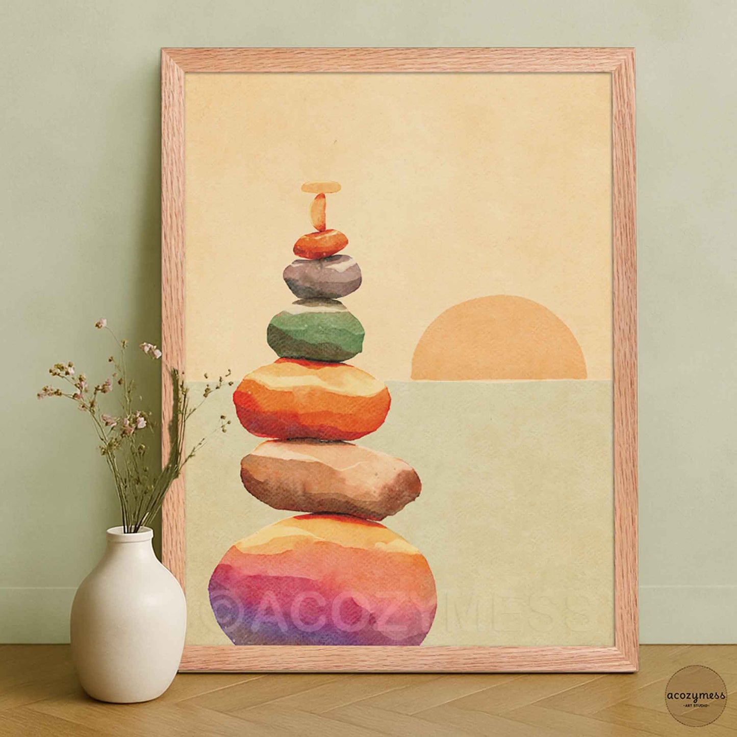 A framed art print featuring a watercolor stack of colorful stones and a sun on a warm beige background, leaning against a sage green wall.