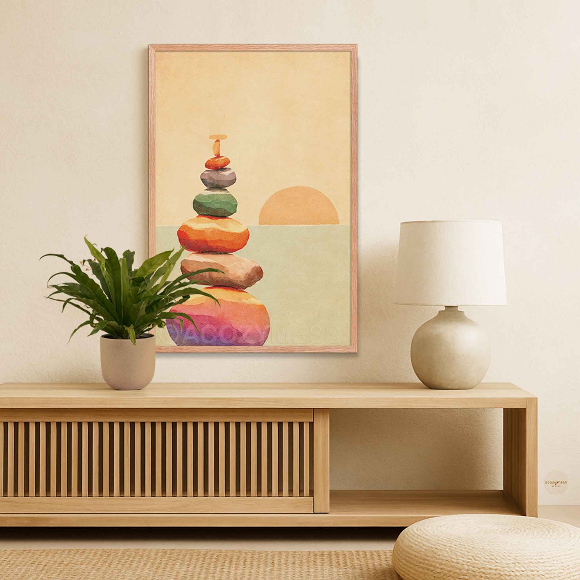 A framed art print featuring a colorful stack of balanced stones against a soft beige and green background, displayed on a light wood console table in a minimalist room setting with a potted plant and a table lamp.