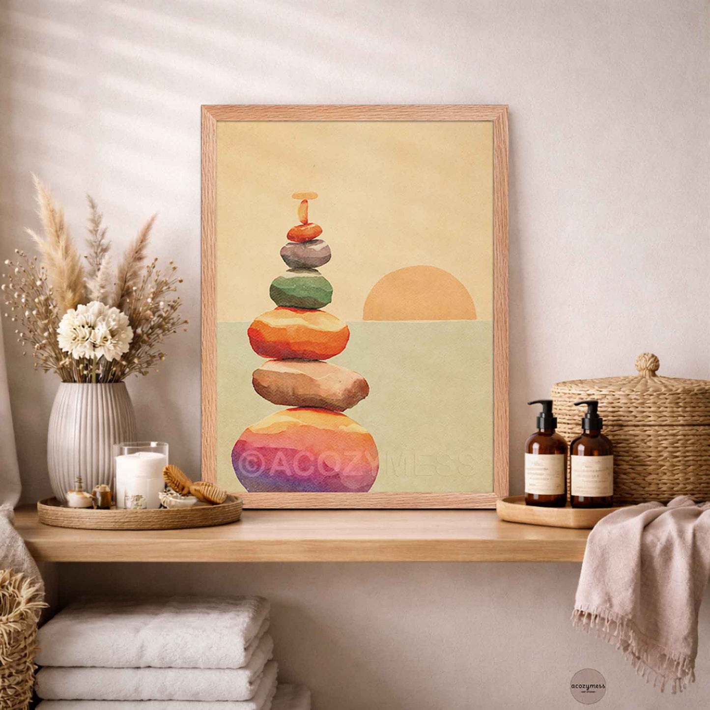 A framed art print featuring a stack of colorful zen stones and a half sun on a beige background, styled on a wooden shelf in a spa-like bathroom setting.