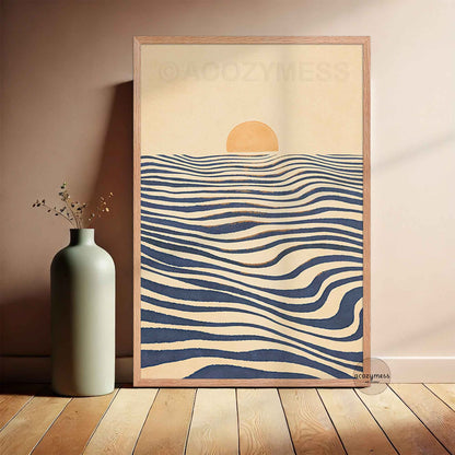 A wooden framed art print leaning against a wall on a wooden floor. The print features a minimalist design with a beige background, a small yellow sun, and wavy navy blue and beige horizontal lines resembling water.