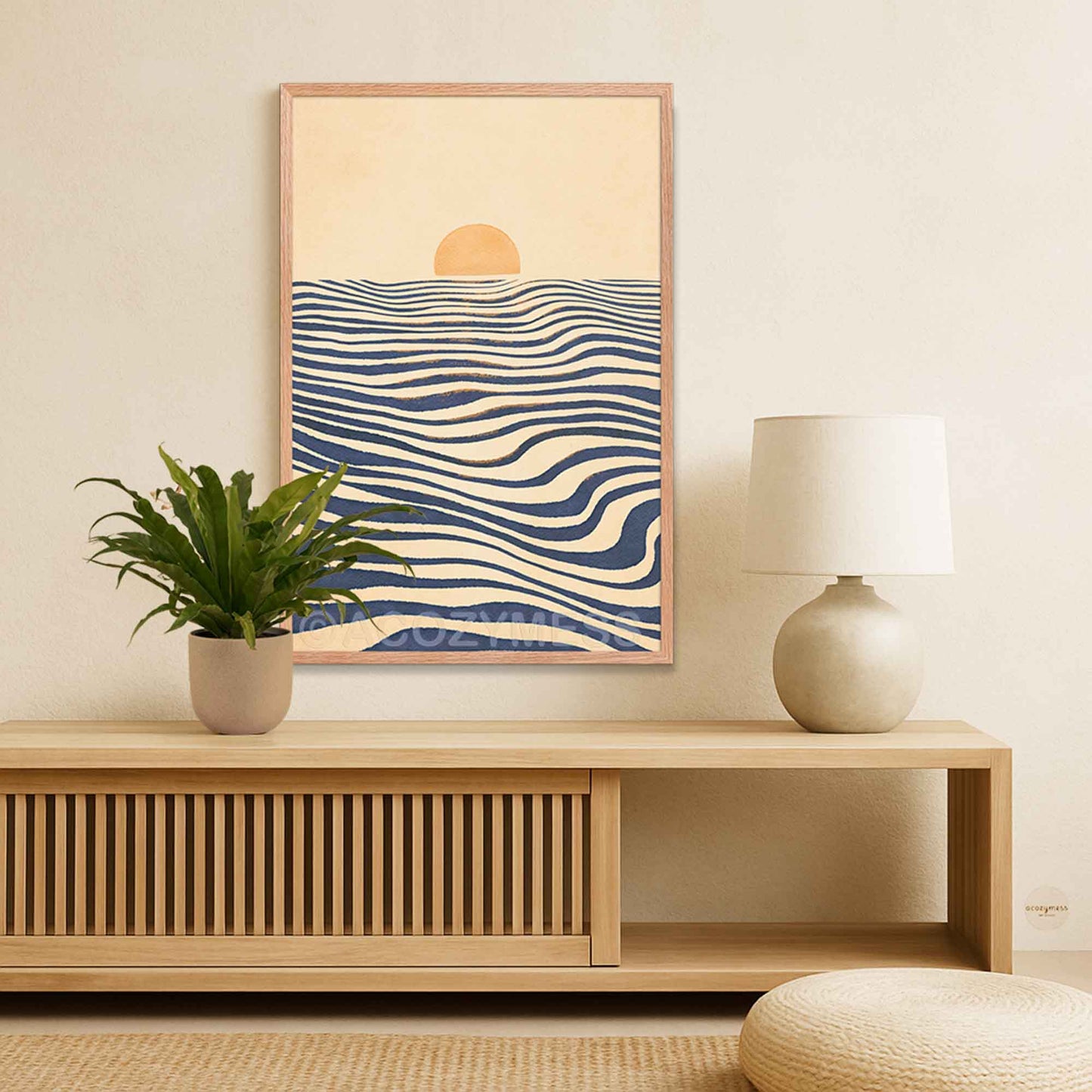 A framed wall art print with a minimalist sun over wavy blue and cream ocean-like lines, displayed above a light wood console table in a styled room.