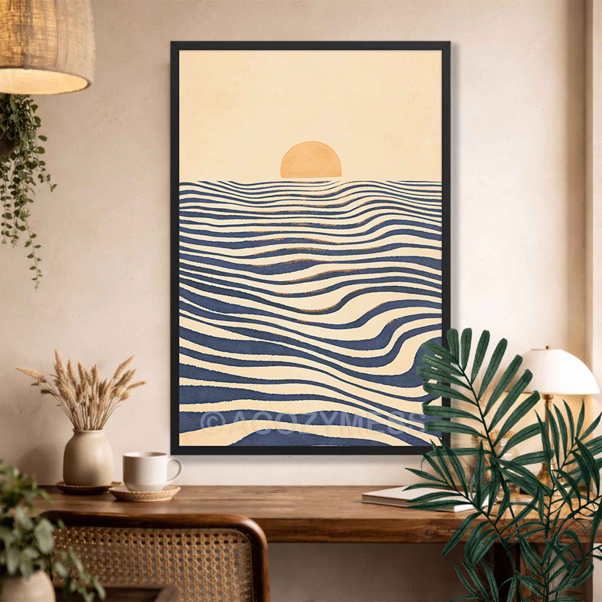 A framed wall art print hangs on a beige wall above a wooden table in a styled interior. The artwork shows a minimalist sunset scene with a pale yellow sun over cream and navy wavy horizontal lines, representing water.