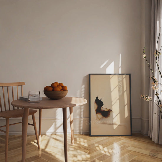Video of a black framed minimalist watercolor cat art poster leaning on the floor beside a wooden dining table in warm sunlight