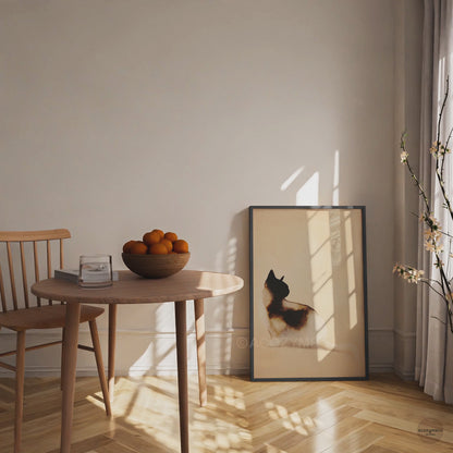 Video of a black framed minimalist watercolor cat art poster leaning on the floor beside a wooden dining table in warm sunlight