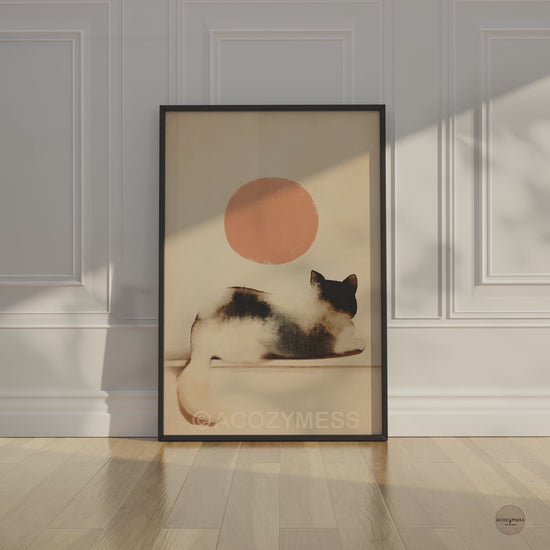 Video of a black framed watercolor cat and peach sun poster leaning against a white paneled wall on a wooden floor in soft sunlight