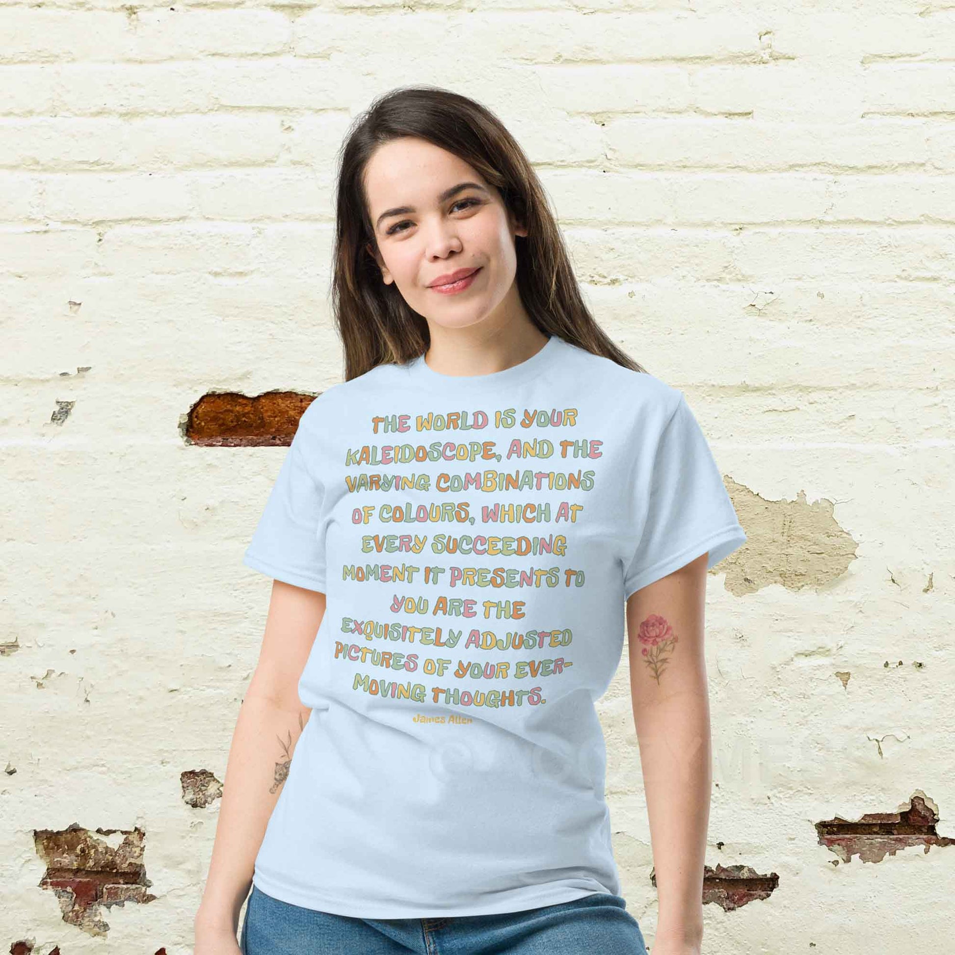 A woman wearing light blue typography T-shirt featuring an inspiring quote by James Allen: The world is your kaleidoscope, and the varying combinations of colors, which at every succeeding moment it presents to you are the exquisitely adjusted pictures of your ever-moving thoughts.