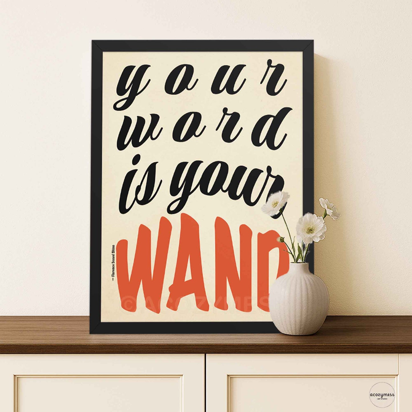 Poster featuring “Your Word Is Your Wand” quote in bold typography, framed in black, on beige background, styled in a minimal modern interior.