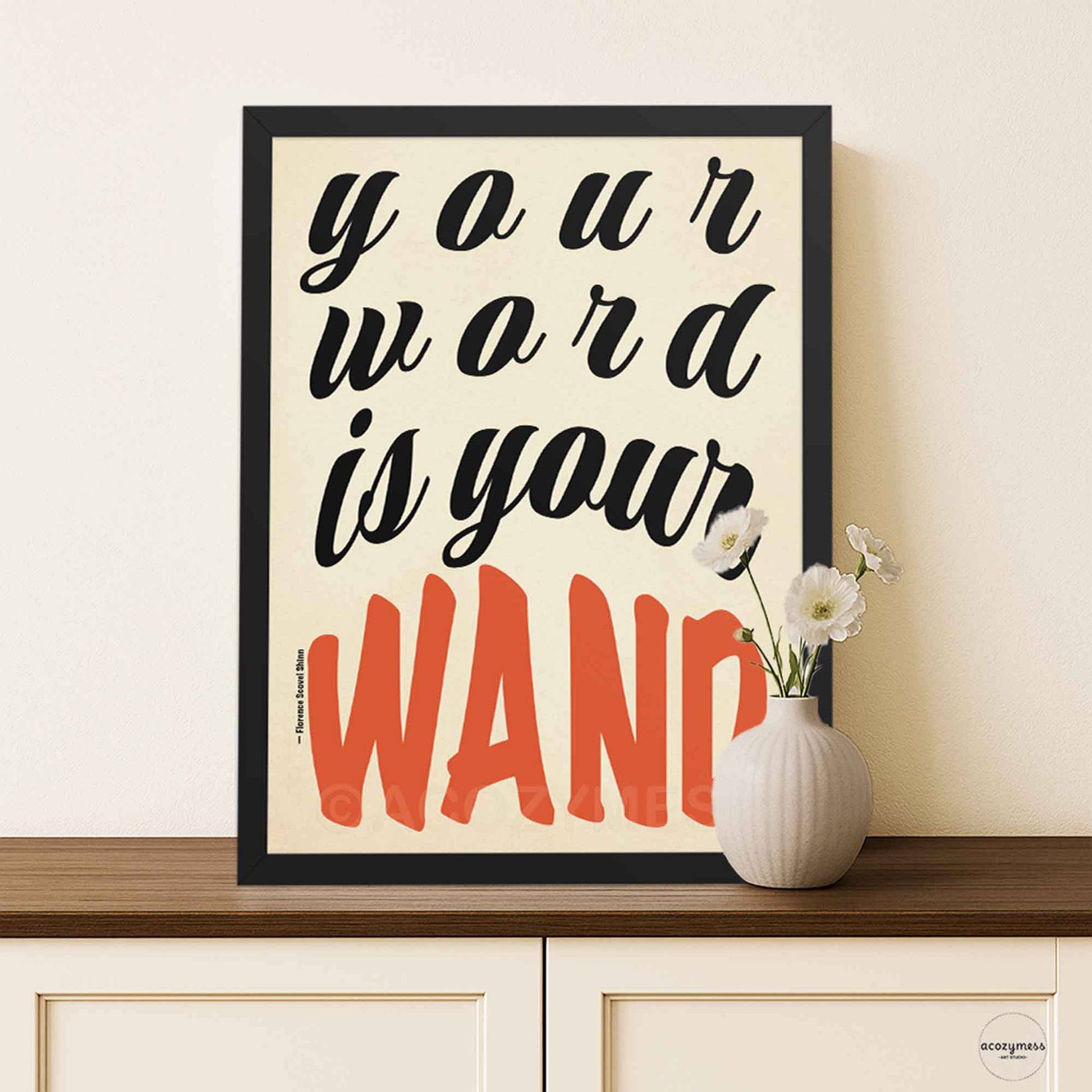 Poster featuring “Your Word Is Your Wand” quote in bold typography, framed in black, on beige background, styled in a minimal modern interior.